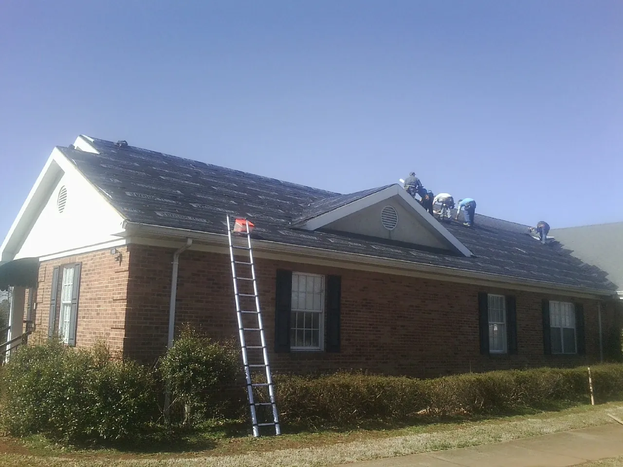 Crew tearing shingles off a brick ranch roof with a ladder set — Emergency Roof Repair work by our crew in Comstock Park