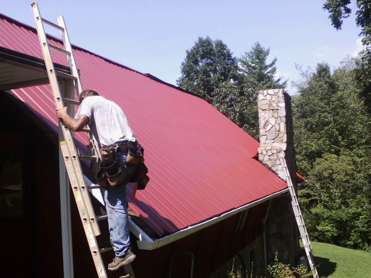 Roofer on a ladder beside a red metal roof and stone chimney — Soffit & Fascia Repair by Roofing Brothers in Comstock Park